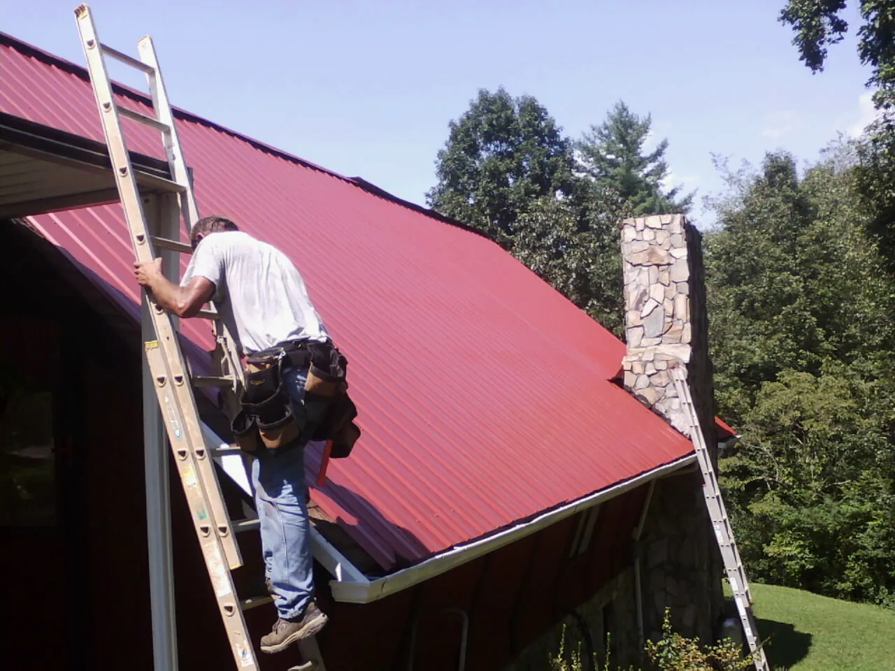 Roofer on a ladder beside a red metal roof and stone chimney — Soffit & Fascia Repair by Roofing Brothers in Bedminster